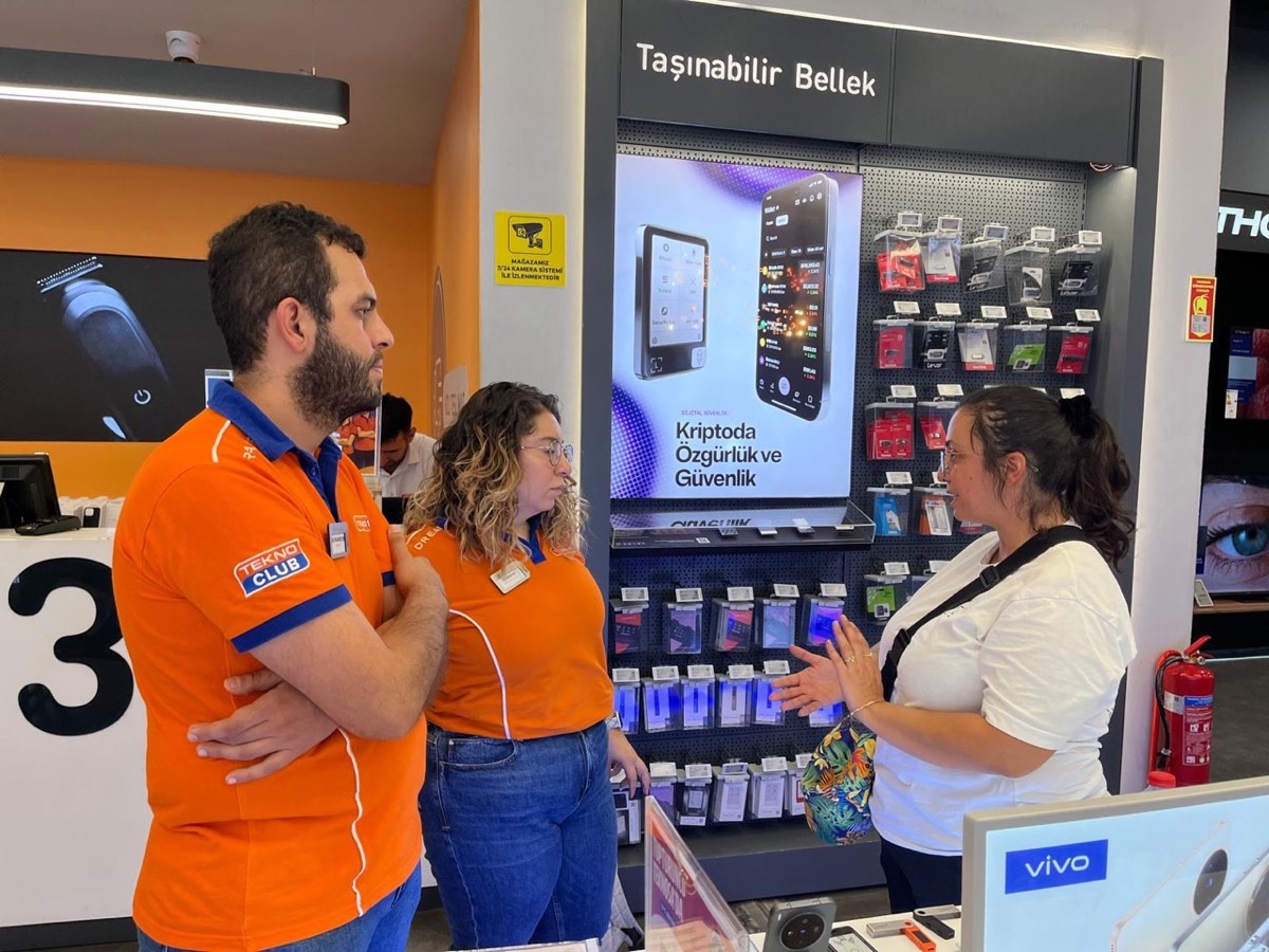 Burcu speaking with Teknosa staff in a store beside a crypto security display